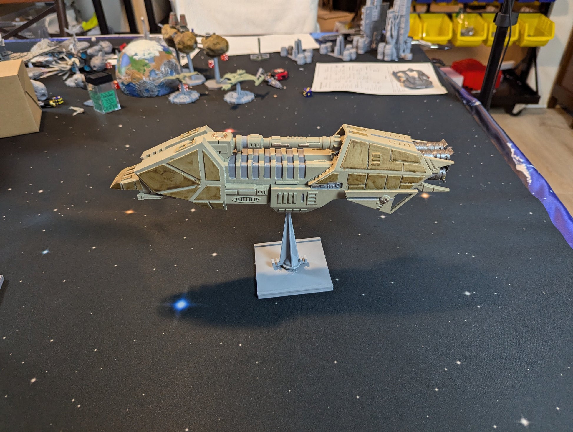 May include: A beige spaceship model with a detailed design, featuring a long, narrow body with multiple compartments and a pointed nose. The spaceship is mounted on a clear plastic stand against a black background with white stars.