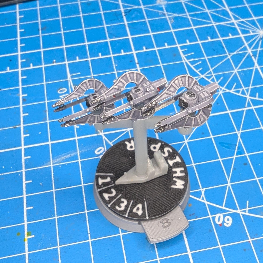 May include: A miniature model of a starship, likely for a tabletop game. The ship is primarily gray with detailed wings and a central body. It is mounted on a black base with white text and numbers, including 'WHIP' and 'R'. The base has a small, angled extension.