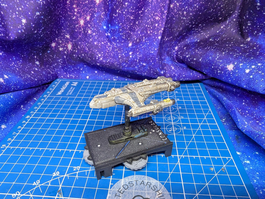 May include: A detailed model of a spaceship, primarily in shades of gray and beige, is displayed on a blue grid surface. The spaceship features intricate detailing and is mounted on a black base with the word 'CONSUL' visible. The background is a vibrant, star-patterned fabric, enhancing the sci-fi theme.
