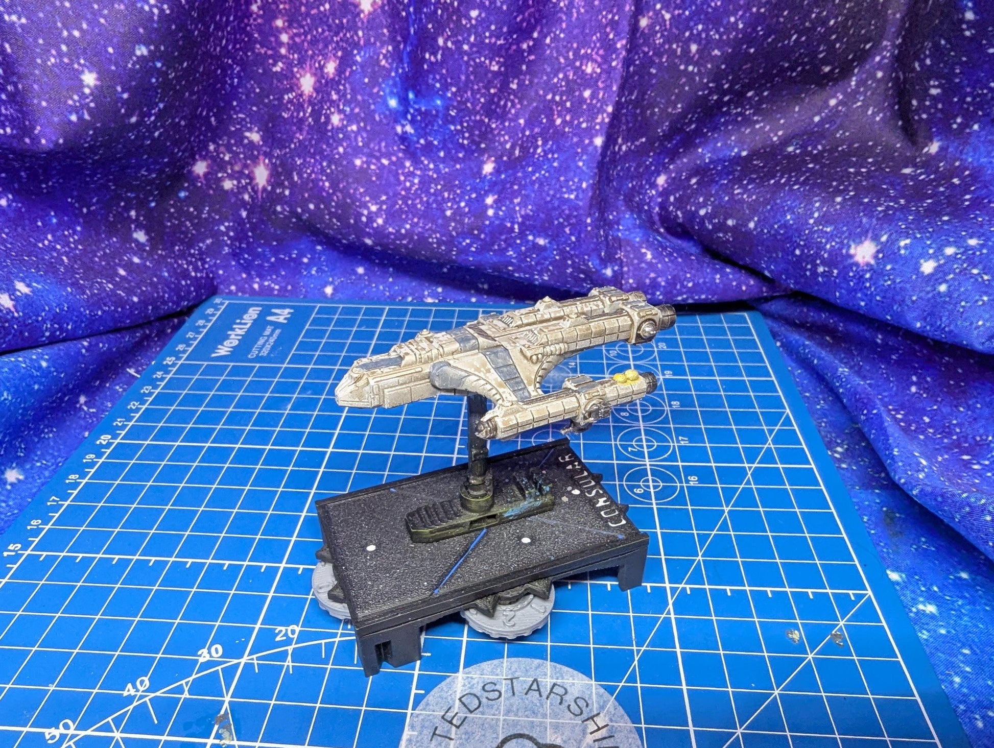 May include: A detailed model of a spaceship, primarily in shades of gray and beige, is displayed on a blue grid surface. The spaceship features intricate detailing and is mounted on a black base with the word 'CONSUL' visible. The background is a vibrant, star-patterned fabric, enhancing the sci-fi theme.