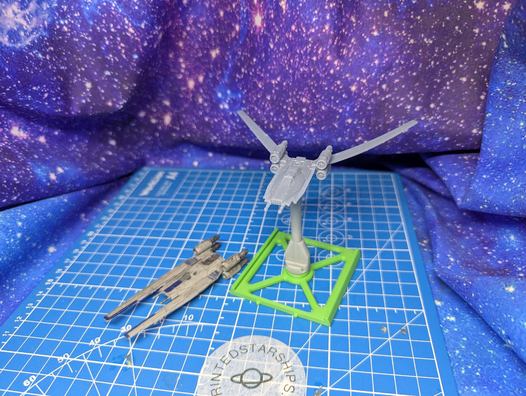 May include: A gray spaceship model with a central body and wings, mounted on a green square base. A second, similar model lies on a blue grid surface. The background is a galaxy-themed fabric. The models appear to be 3D printed.