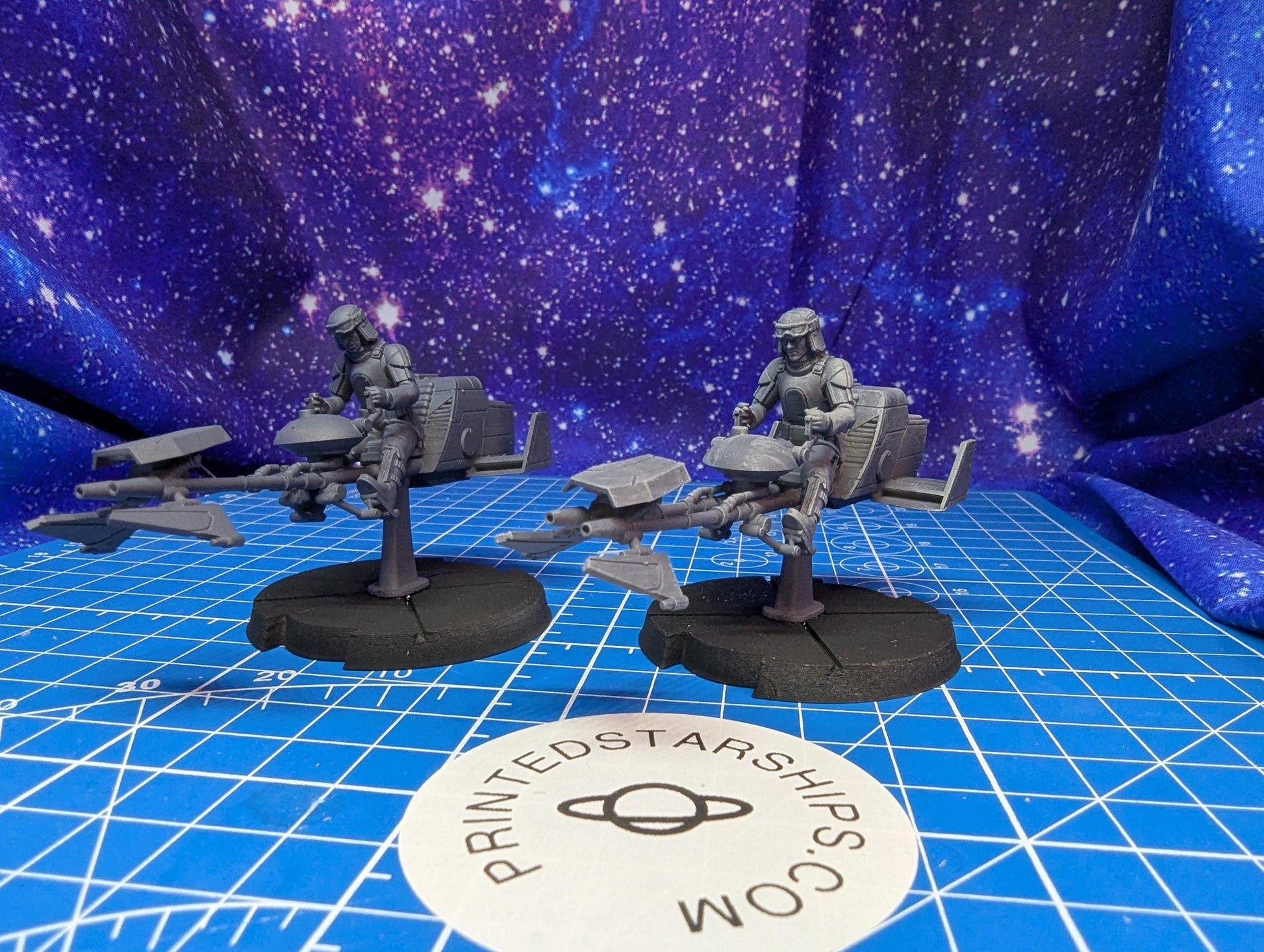 May include: Two gray 3D printed Star Wars speeder bikes with figures. The speeder bikes are on black bases with a printed logo that says 'PRINTEDSTARSHIPS.COM'.