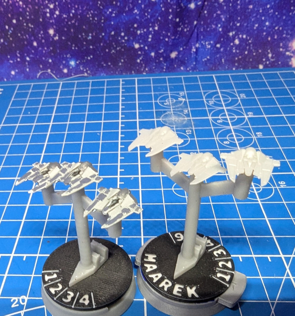 May include: Two sets of miniature spaceships on stands. The spaceships are painted in a light gray color. The stands are black with white numbers and text. The text on the stands reads '1 2 3 4' and 'MAAREK 9 7/2/1'.