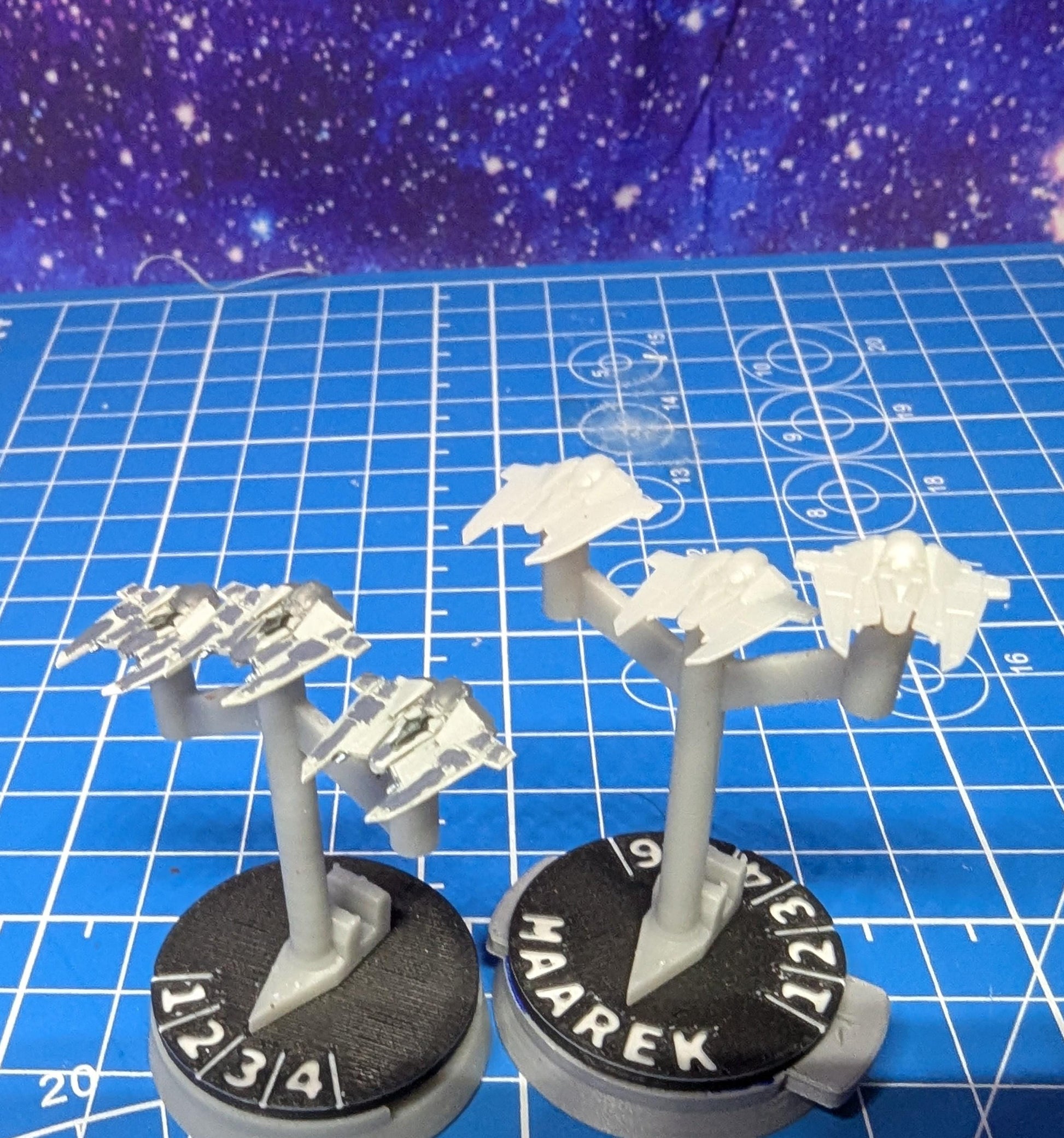 May include: Two sets of miniature spaceships on stands. The spaceships are painted in a light gray color. The stands are black with white numbers and text. The text on the stands reads '1 2 3 4' and 'MAAREK 9 7/2/1'.