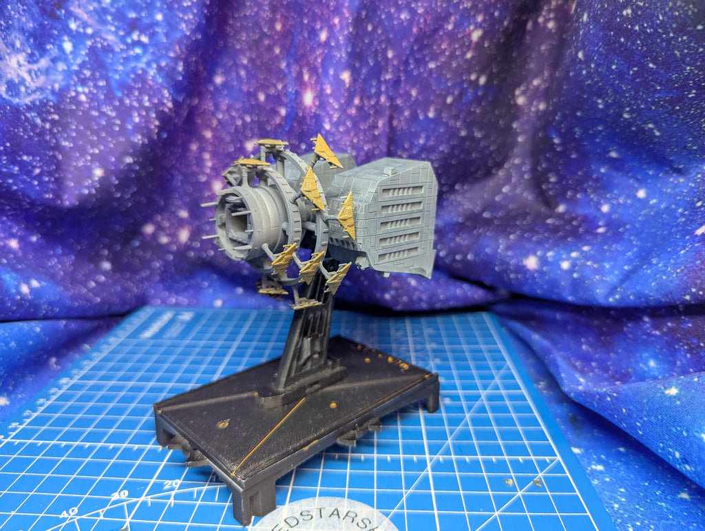 May include: A detailed model of a futuristic engine. The engine is primarily gray with gold accents and a rectangular component. It is mounted on a black base with a grid pattern, set against a blue and purple galaxy backdrop.
