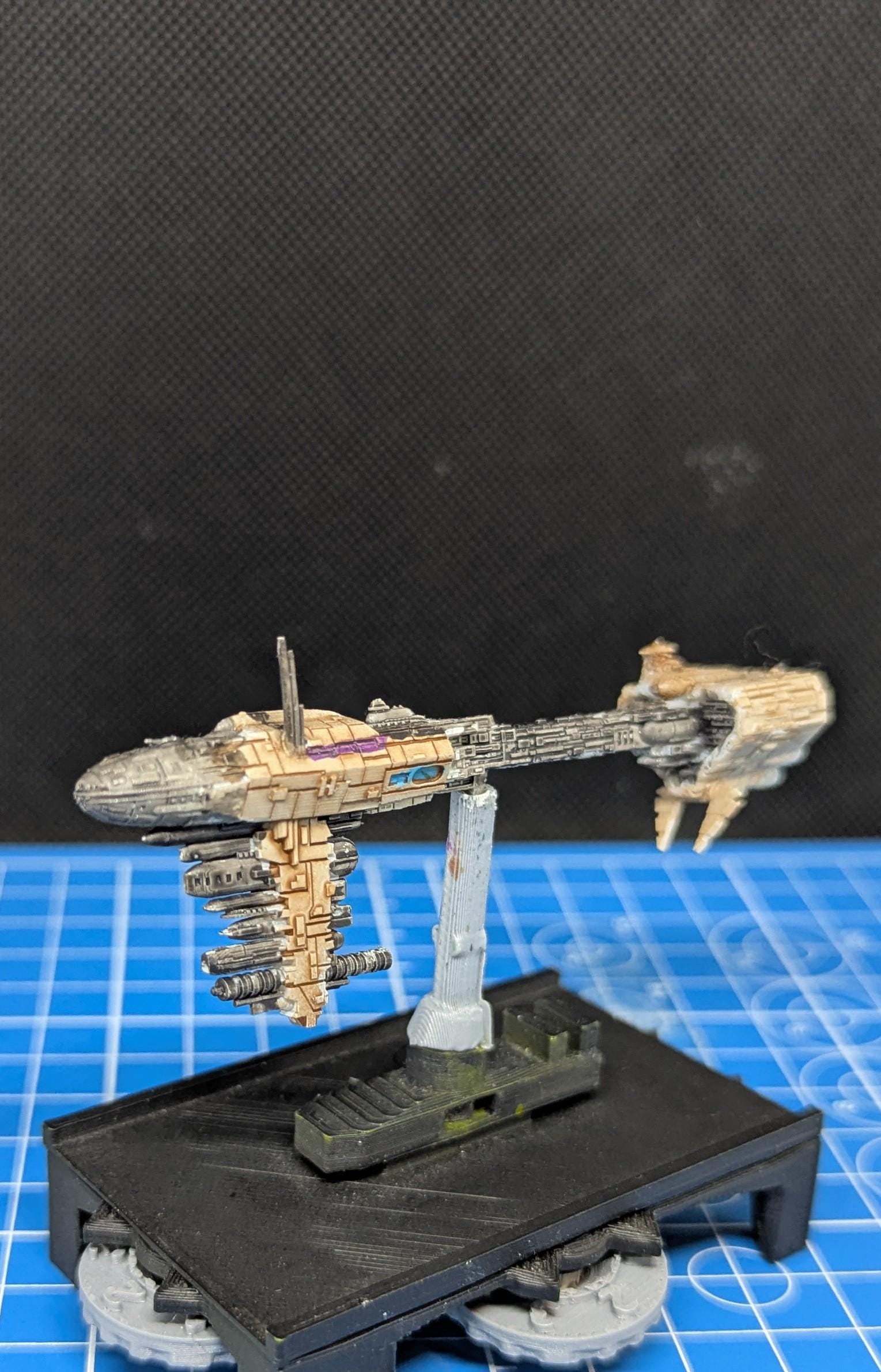 May include: A beige and gray spaceship model with a long, thin body and a large, round cockpit. The spaceship is mounted on a black base with a gray stand. The base has a textured surface and is decorated with green paint. The spaceship is a miniature model of a fictional spacecraft.