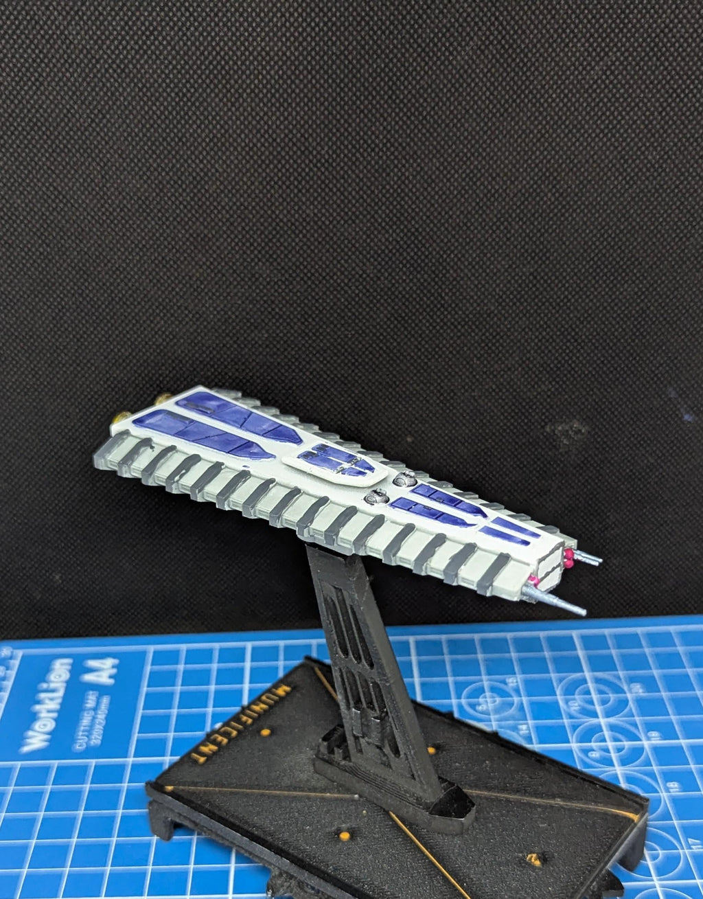 May include: A white and blue spaceship model with gray details. The spaceship is on a black display stand. The stand is on a blue grid surface. The text 'MULTIFLEX' is visible on the stand.