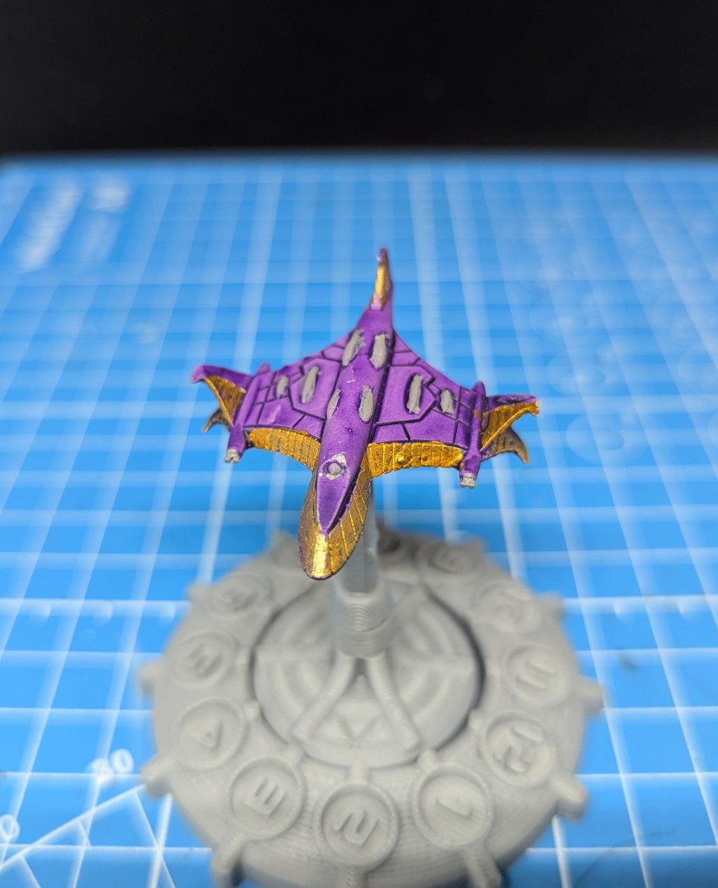 May include: A purple and gold spaceship model on a gray base with a circular design. The spaceship has a pointed nose and wings that are angled upward.