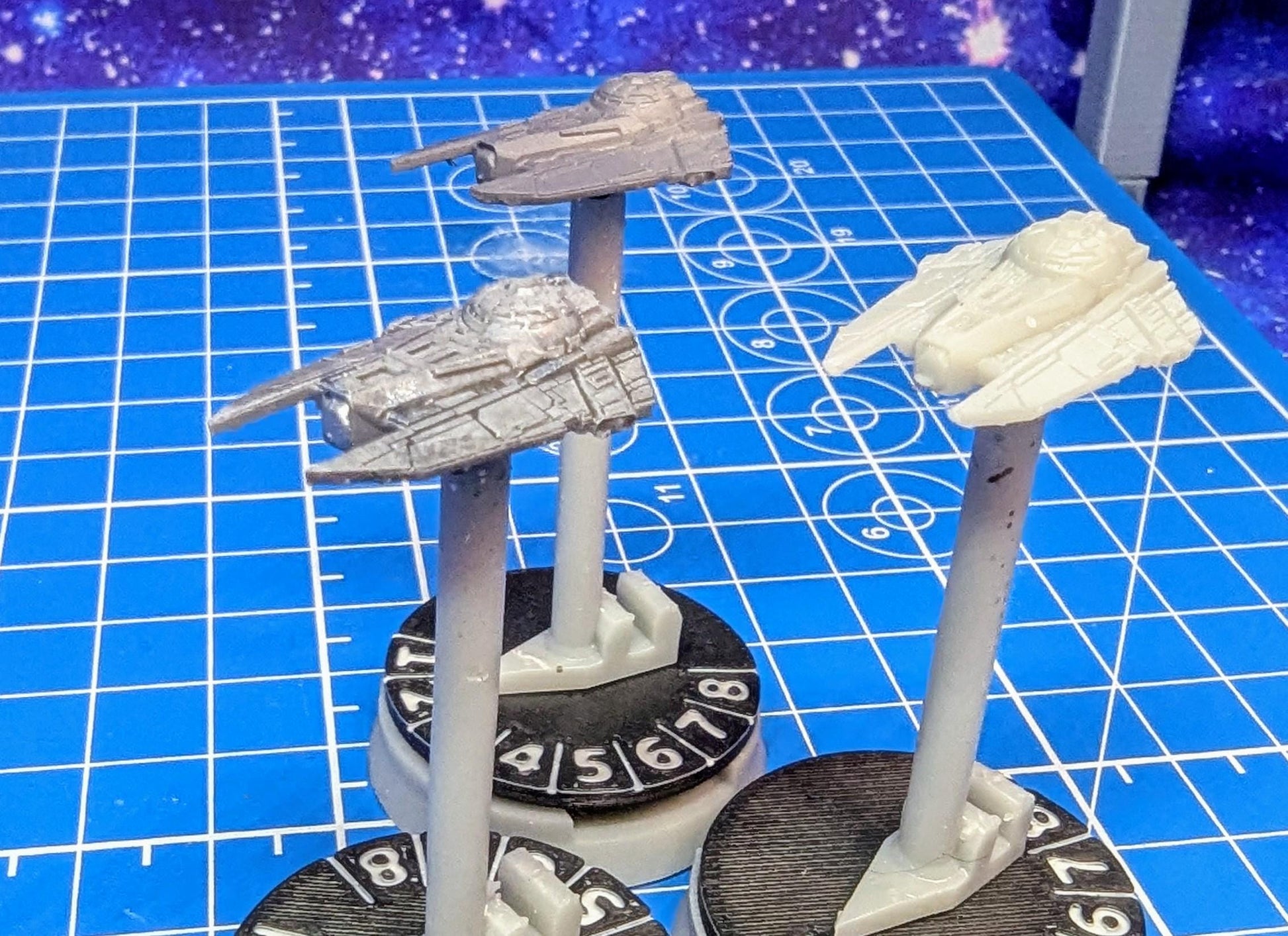 May include: Three miniature spaceships painted in gray and white on black bases with white numbers 1 through 8. The spaceships are on a blue grid background.
