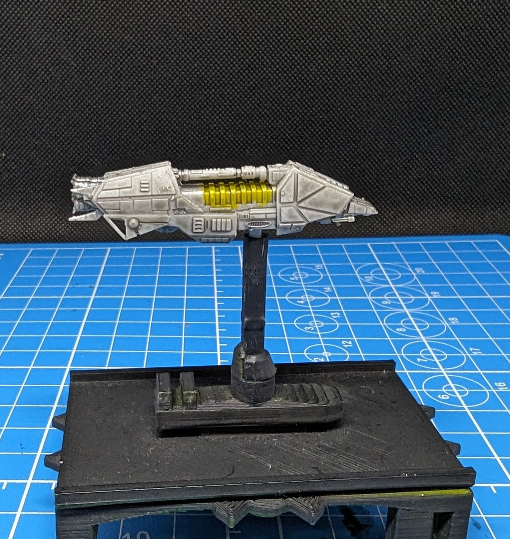 May include: A gray and white spaceship model with yellow details. The spaceship is mounted on a black stand and is displayed on a blue grid surface.