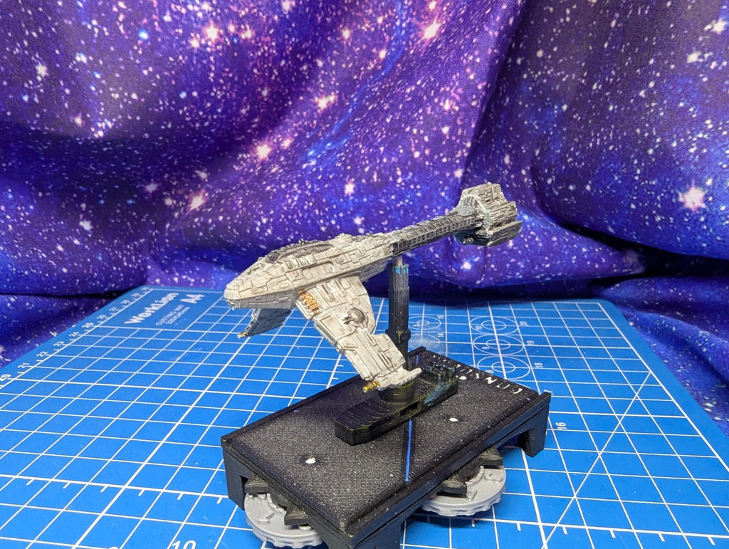 May include: A detailed model of a spaceship, primarily gray with some tan accents, is displayed. The spaceship is mounted on a black base with a clear support. The base has a circular design with visible gears. The background features a starry, purple and blue fabric.