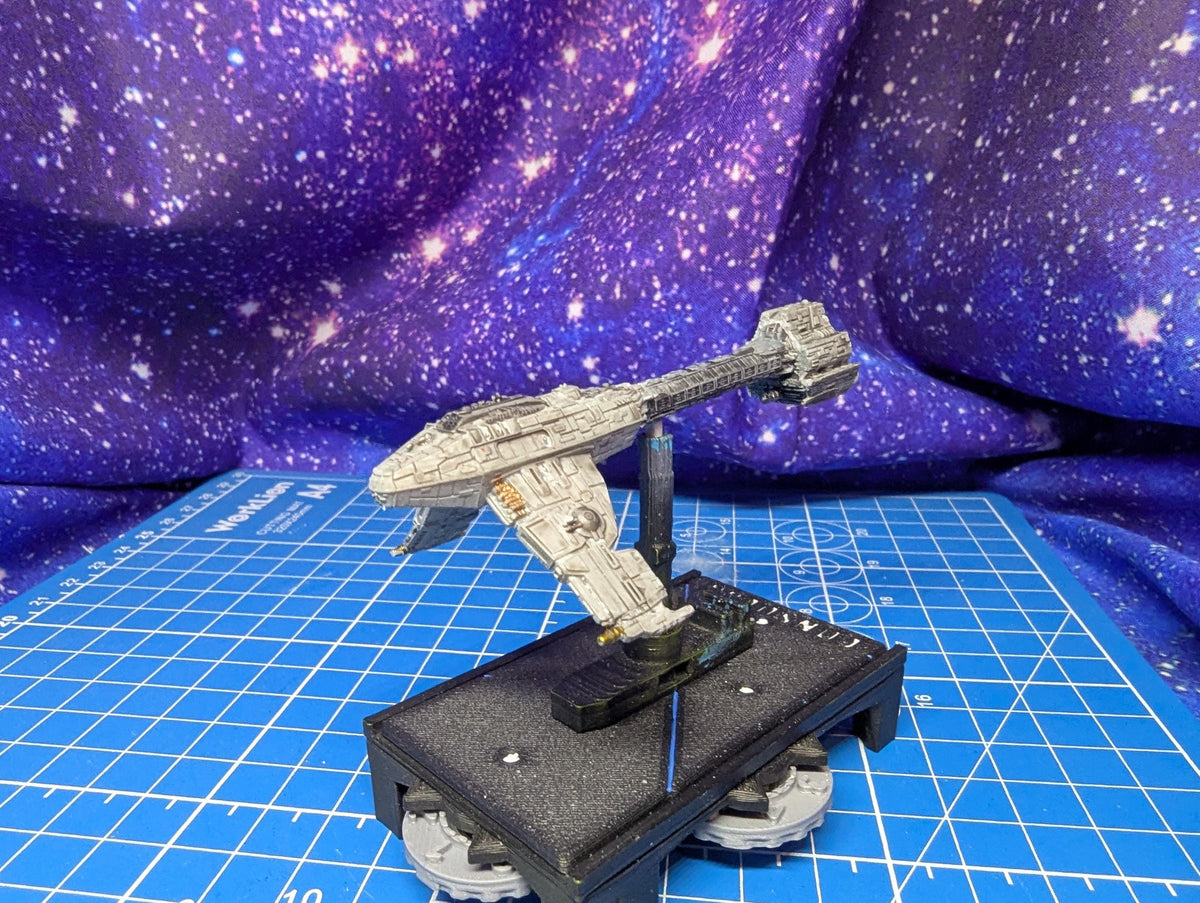 May include: A detailed model of a spaceship, primarily gray with some tan accents, is displayed. The spaceship is mounted on a black base with a clear support. The base has a circular design with visible gears. The background features a starry, purple and blue fabric.