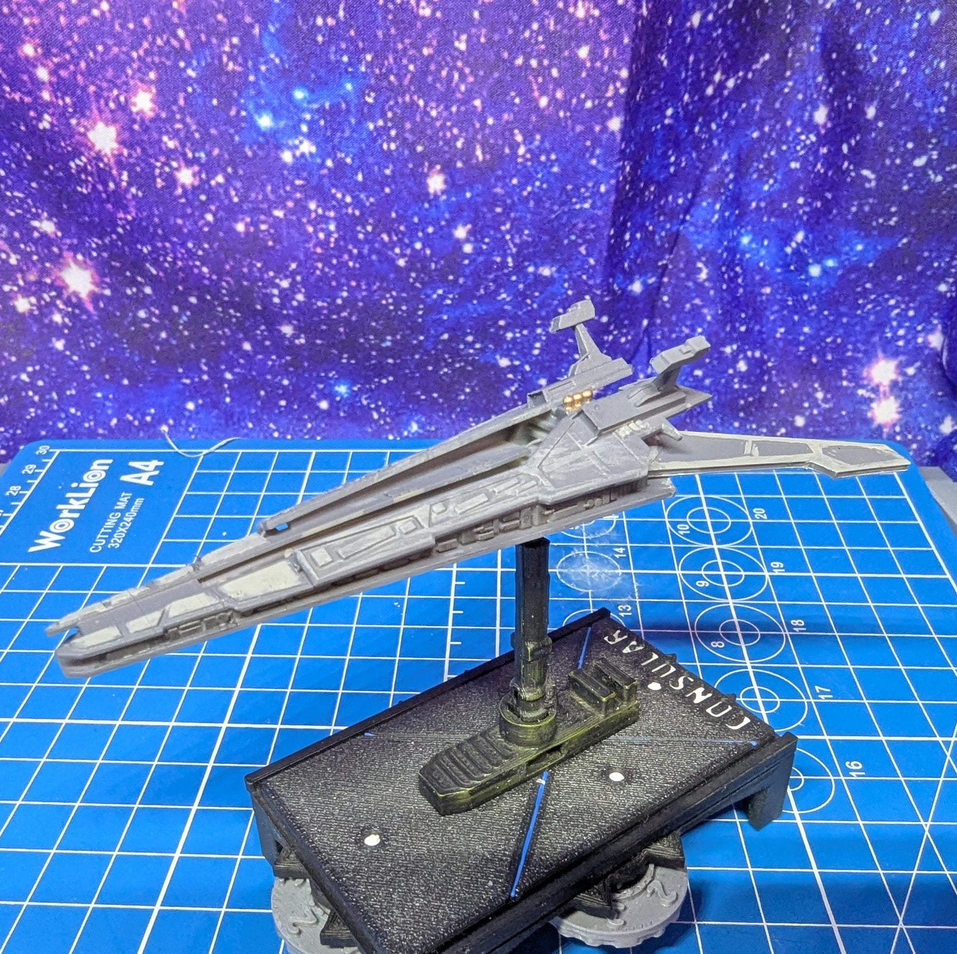 May include: A gray spaceship model on a black base with a blue and gray grid background. The spaceship has a long, thin body with a pointed nose and wings. The base has a circular design with the word 'CONSULAR' printed on it.